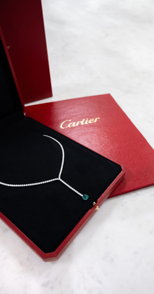 Cartier diamond necklace with a single emerald stone in an open red Cartier box, with a Cartier certificate folder. This showcases the designer jewelry items that Diamond Banc in Boca Raton is interesting purchasing. 