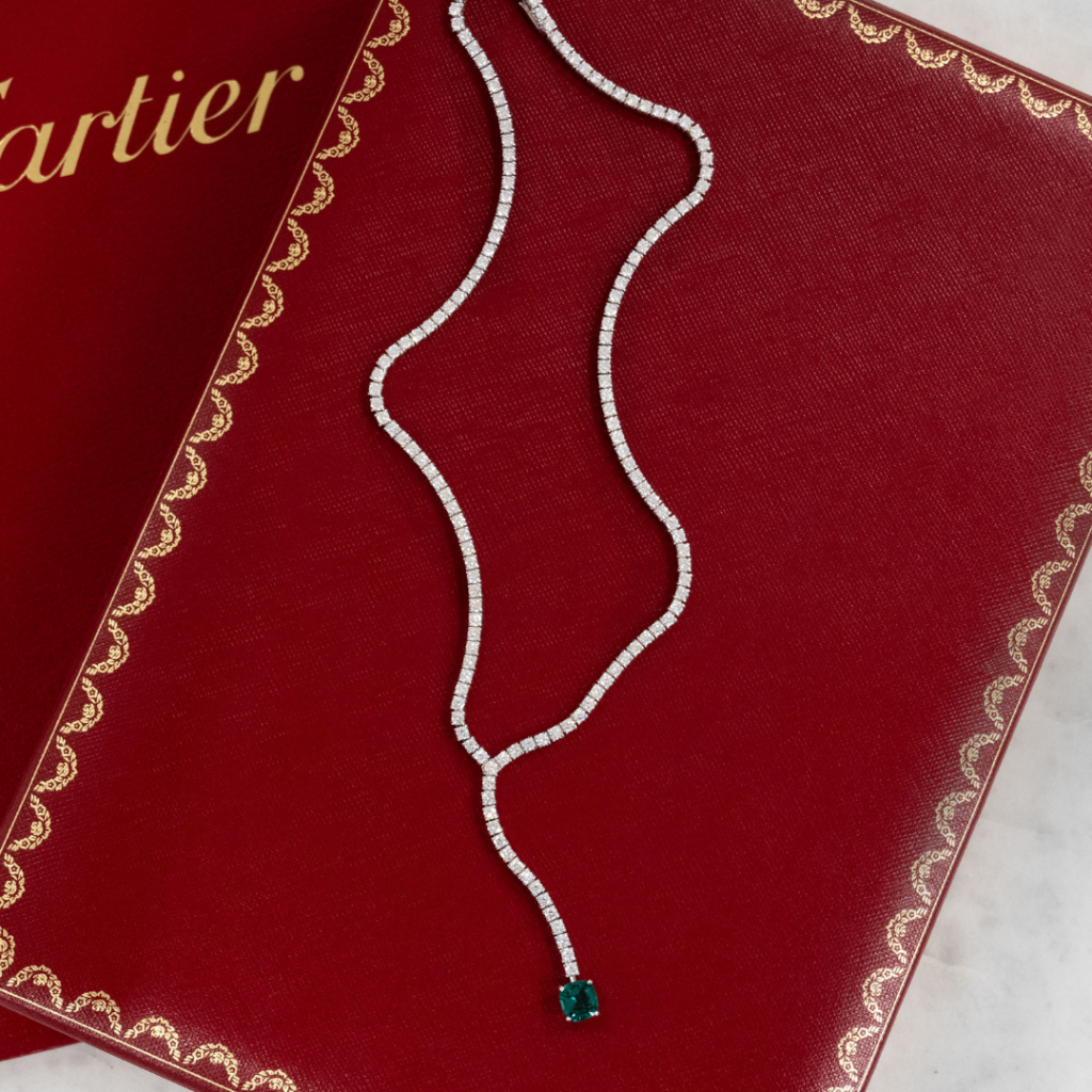 Featuring a Cartier diamond necklace with a vibrant green emerald pendant on a Cartier box, positioned above a Cartier certificate folder,  Diamond Banc is the premier destination for clients interested in selling their luxury jewelry to a reputable jewelry buyer in Sarasota.