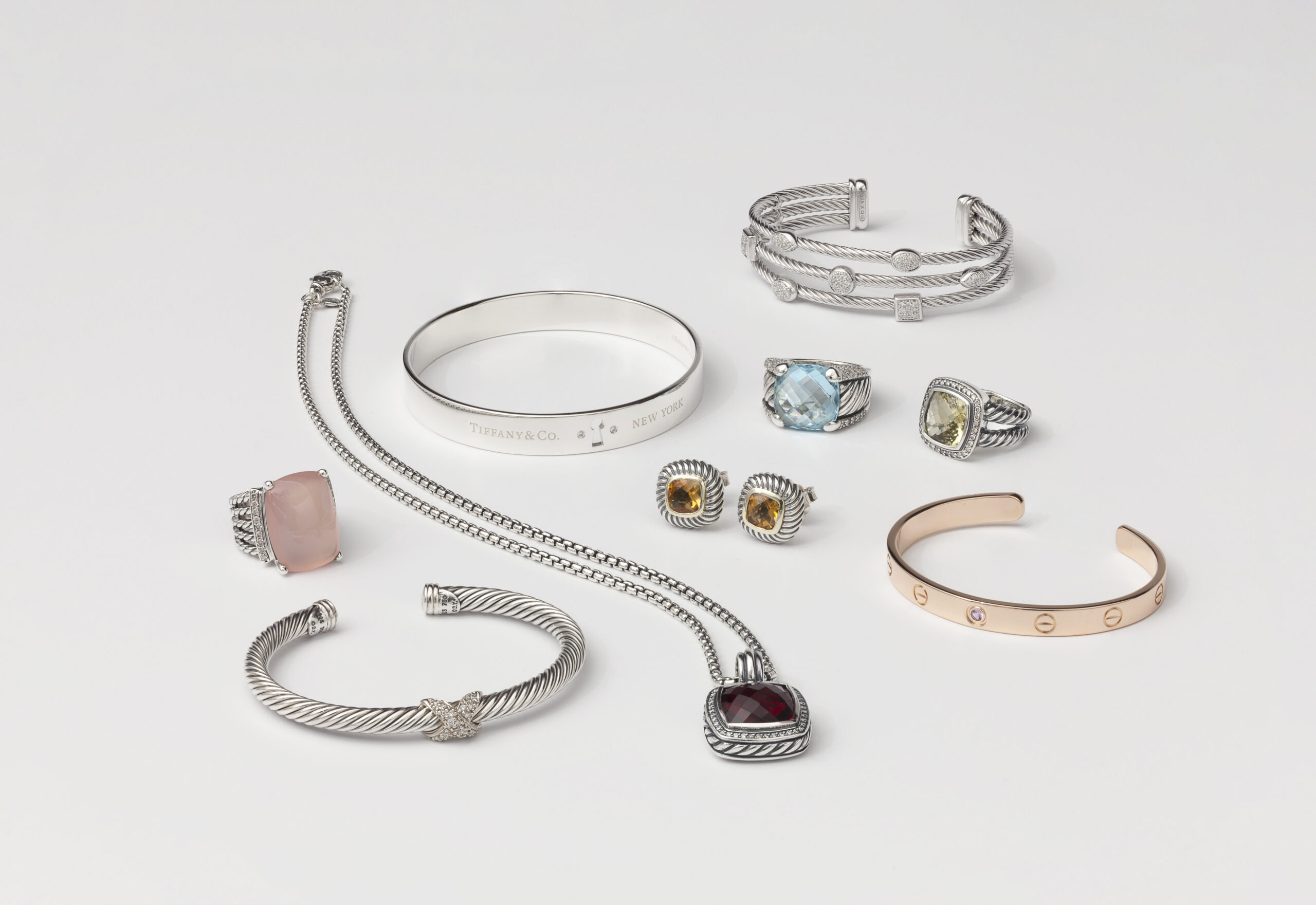 Collection of luxury designer jewelry, including Tiffany & Co. and Cartier bracelets, gemstone rings, cable-inspired cuffs, and pendant necklaces in silver and gold.